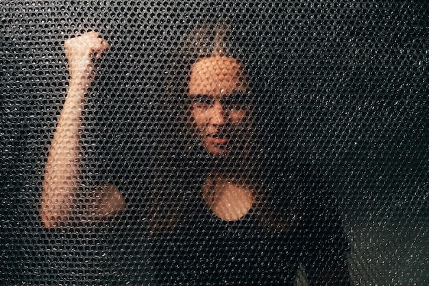 Woman protest Female rigths empowerment Gender equality Textured portrait of annoyed angry mad feminist activist lady in black knocking on plastic bubble wrap wall isolated in darkness