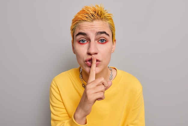 Photo woman punk with trendy yellow hairstyle bright colorful makeup makes silence gesture shushing indoor wears casual jumper poses against grey. hush be quiet please