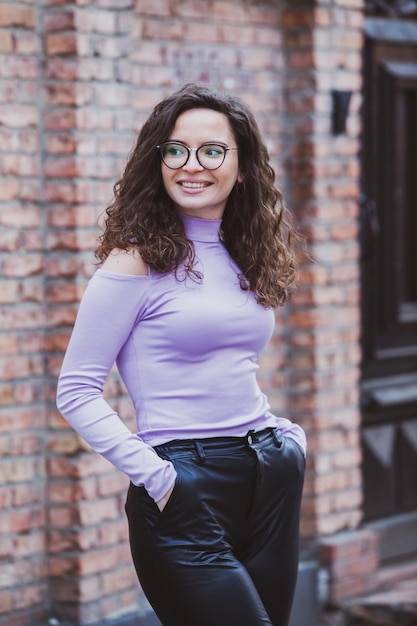 A woman in a purple top and black pants stands in front of a brick wall.