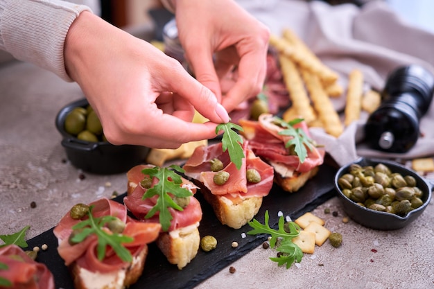 Woman puts arugula leaves to prosciutto ham bruschetta with traditional antipasto meat plate on background