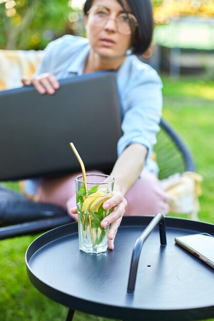 Woman puts down the laptop and takes lemonade sitting on chair at backyard