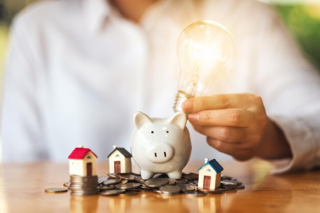 A woman putting light bulb over a piggy bank with house models and pile of coins on the table for saving money concept