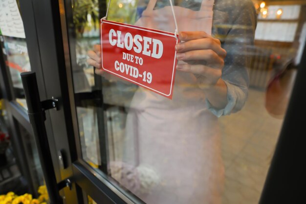 Woman putting red sign with words Closed Due To Covid19 onto glass door closeup