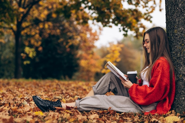 Woman reading in an autumn park and drinking coffee under the tree