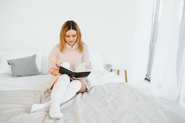 A woman reading a book and smiling as she sits in bed. The alarm clock on the desk beside her.