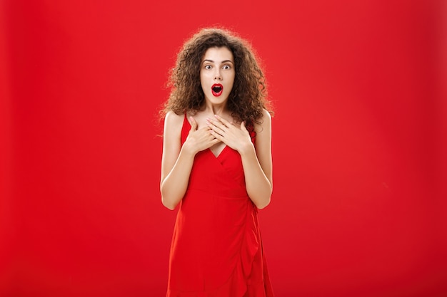 Woman receiving awesome news being impressed and amazed in red evening dress gasping saying wow holding hands on brest standing astonished and excited over red background gossiping. Copy space