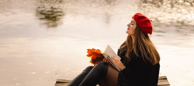 Woman in a red beret reading book on wooden pontoon Autumn season Female with trendy clothes relaxing near water reading book and drink coffee Autumn mood Autumn concept Autumn day