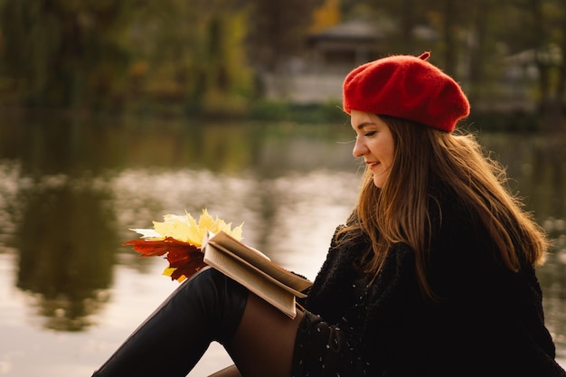 Woman in a red beret reading book on wooden pontoon Autumn season Female with trendy clothes relaxing near water reading book and drink coffee Autumn mood Autumn concept Autumn day