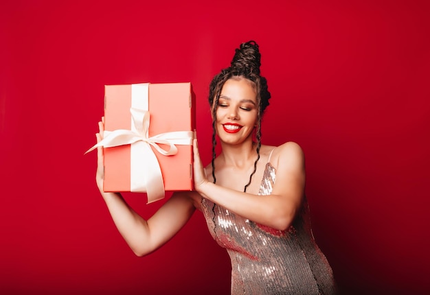 A woman in a red Christmas hat with dreadlocks holds a gift on a red background Isolate a woman in a smart dress new year christmas