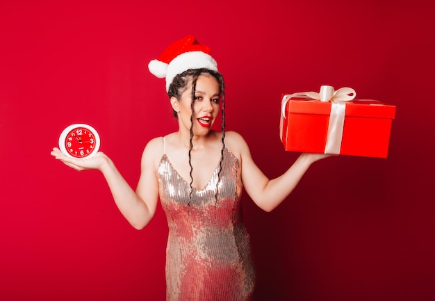 A woman in a red Christmas hat with dreadlocks holds a gift on a red background Isolate a woman in a smart dress new year christmas