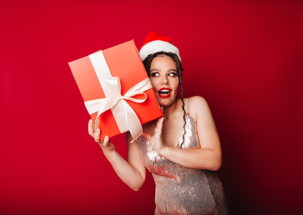 A woman in a red Christmas hat with dreadlocks holds a gift on a red background Isolate a woman in a smart dress new year christmas