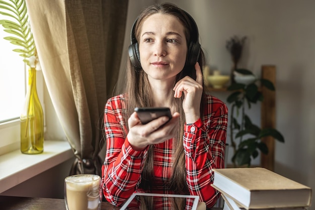 A woman in a red dress is listening audiobook wearing headphones with phone by the window The concept of modern technological education and the study of books with joy