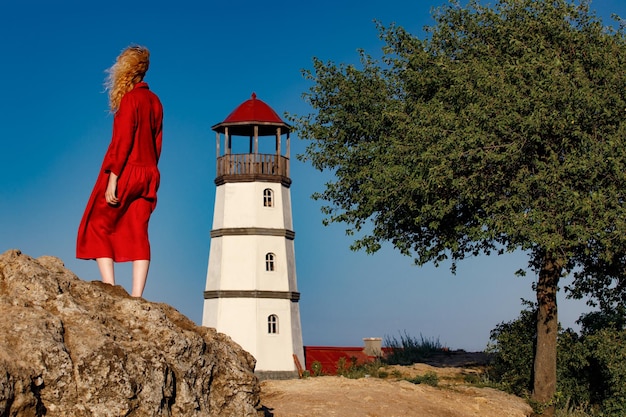 A woman in a red dress is standing on the beach near the lighthouse at sunrise.