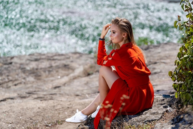 Photo a woman in a red dress sits on a rock by the water she looks sad and is wearing a watch