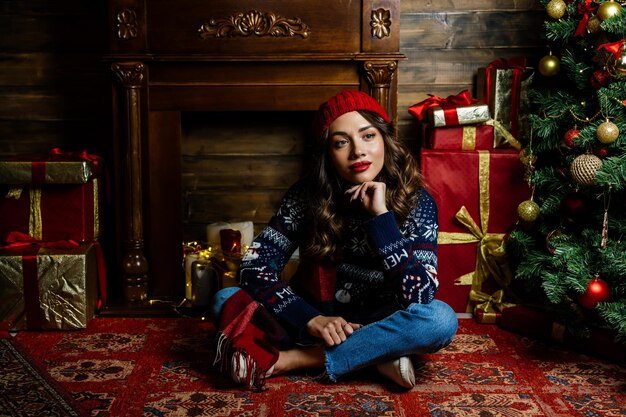 A woman in a red hat and scarf thought A beautiful brunette sits under a Christmas tree against the backdrop of New Year's decorations