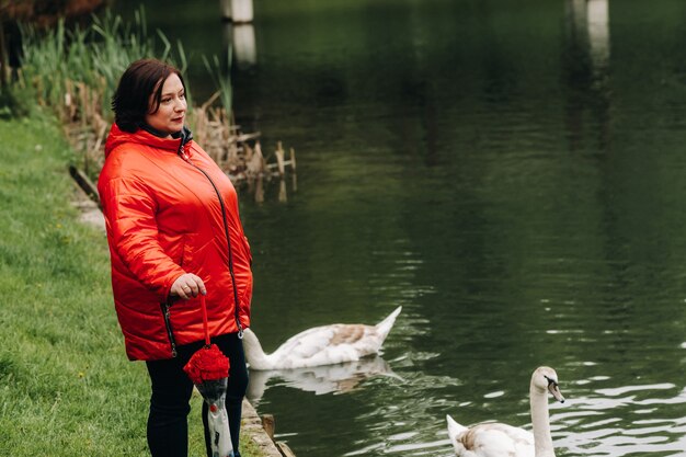 A woman in a red jacket and carrying an umbrella walks through a summer park near a lake with swans