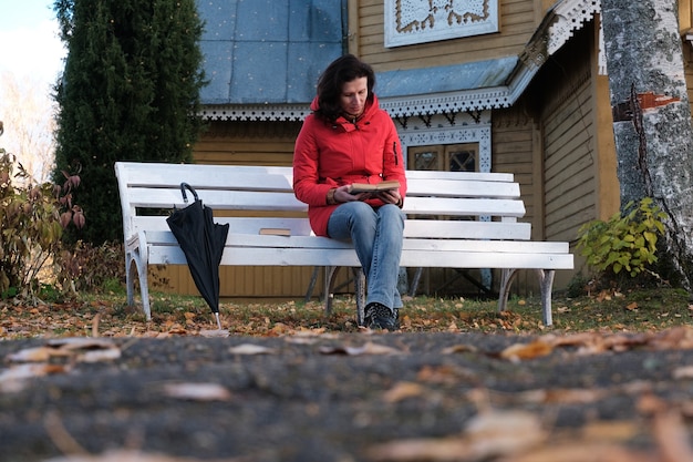 A Woman in a Red Jacket Is Sitting on a White Bench in the Park and Reading A Paper Book.