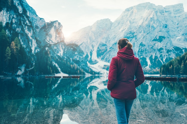 Woman in red jacket is standing on the coast of Braies lake at sunrise in autumn. Dolomites, Italy.