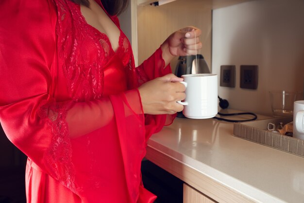 A woman in a red silk nightgown and luxurious robes making hot tea in the kitchen at night