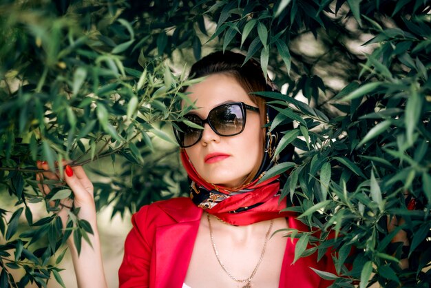 Woman in a red suit and headscarf wearing sunglasses against green leaves