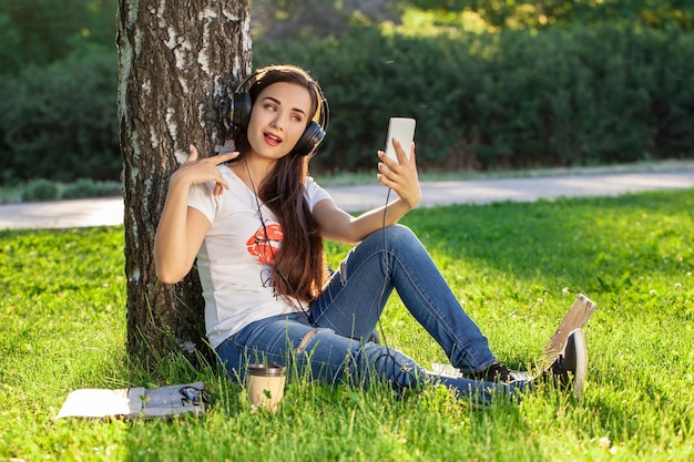 Woman relax with headphones listening to music sitting on grass in park. Young woman with headphones doing selfie on her smartphone