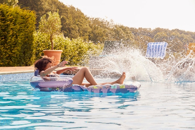 Woman Relaxing Floating On Inflatable In Swimming Pool As Teenage Son Jumps In On Summer Vacation