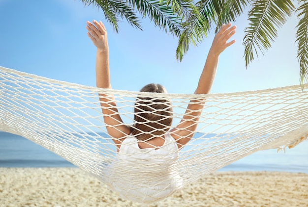 Woman relaxing in hammock under green palm leaves on sunlit beach