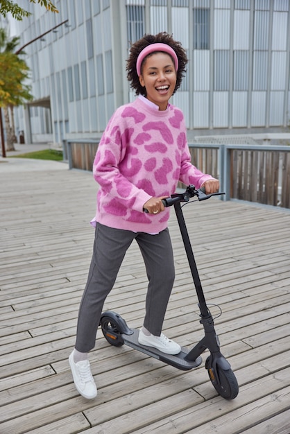 woman rides electric scooter against urban background enjoys good day dressed in jumper trousers and sneakers has fun outdoors during sunny day