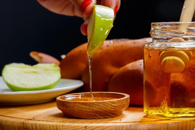 A woman's hand dips a piece of apple in honey in honor of the celebration of Rosh Hashanah next to honey and challah