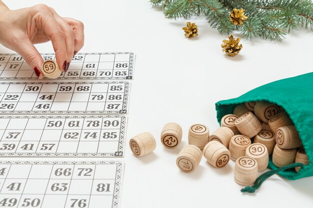 Woman's hand holding a barrel for a game in lotto. Wooden lotto barrels with a green bag and game cards. Christmas fir tree branches on the background. Board game lotto