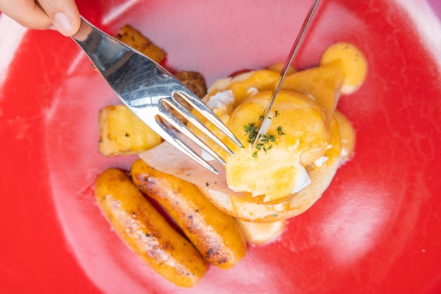 Woman's hand holding a fork and eating Breakfast Egg Benedict fruits such as watermelon papaya melon passion fruit orange juice and coffee placed on a gray placemat