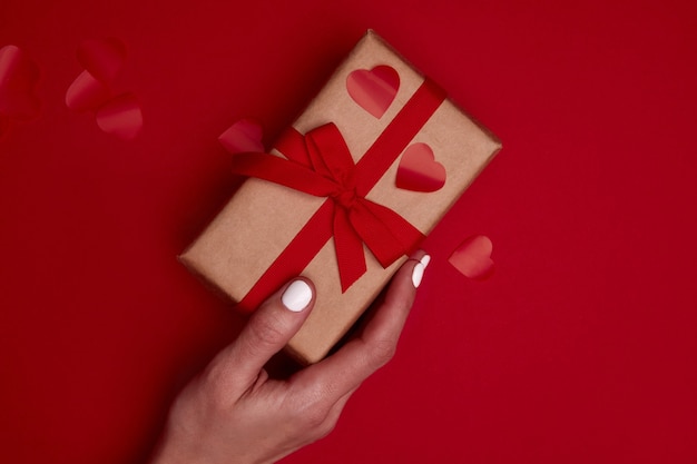 Woman's hand holding Valentine gift box wraped in craft paper with ribbon bow and red hearts