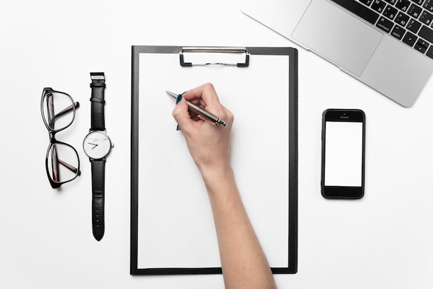 A woman's hand holds a pen and writes on a clean sheet of paper. White office desk with laptop, phone and supplies.