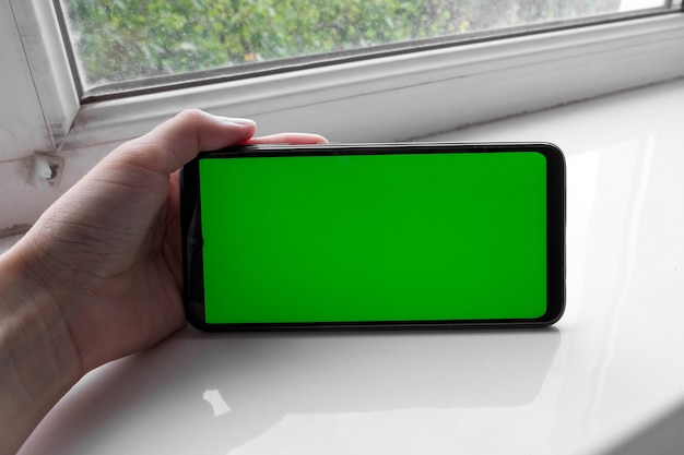 The woman's hand holds the smartphone in a horizontal position with a green screen on a window. Chroma key. Mock up.