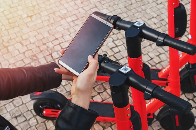 Woman's hands close up holding a phone near the modern city electric scooter