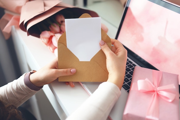 Woman's hands holding a letter in craft envelope. Pink background, valentine's day concept. Tulips flower and pink gift box in background. Womens home desk.