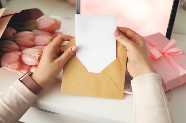 Woman's hands holding a letter in craft envelope. Pink background, valentine's day concept. Tulips flower and pink gift box in background. Womens home desk.