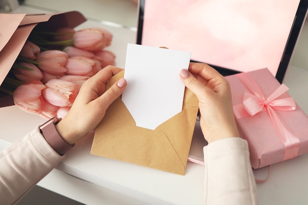 Woman's hands holding a letter in craft envelope. Pink background, valentine's day concept. Tulips flower and pink gift box in background. Womens home desk.