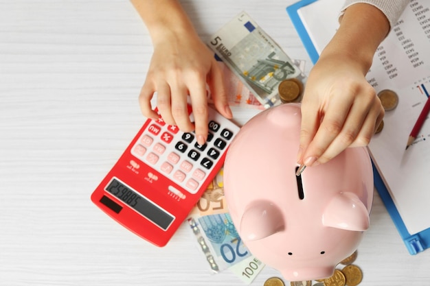 Woman's hands putting euro coin into a piggy bank and counting with calculator on the table Financial savings concept