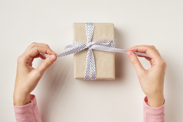 Woman's hands untie bow on gift box on white background, top view