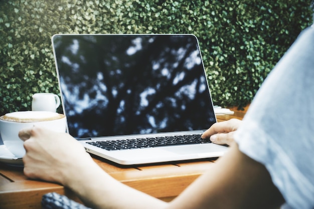 Woman's hands using laptop and drinking cappuccino Green bush in the background Mock up