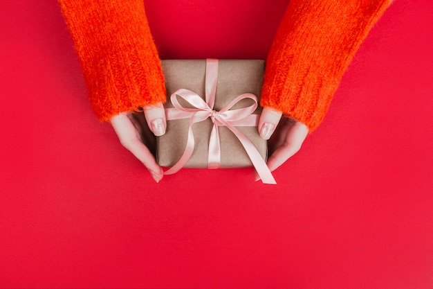 Woman's hands in warm knitted sweater with manicure hold gift box wrapped with craft paper and pink ribbon on red.