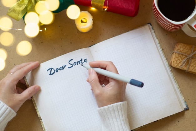 Woman's hands in white sweater writing letter to Santa in notebook, selective focus, bokeh lights, atmospheric photo. Christmas shopping and gifts concept.