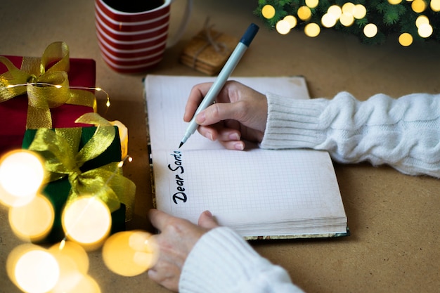 Woman's hands in white sweater writing letter to Santa in notebook, selective focus, bokeh lights, atmospheric photo. Christmas shopping and gifts concept.