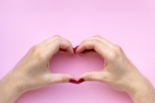 Woman's hands with red color nails making heart symbol on pink wall