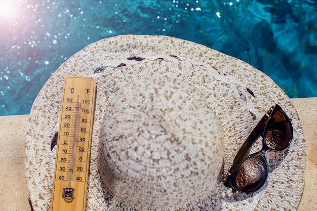 Woman's hat, thermometer and sunglasses lying beside the pool. Hot weather, summer and holiday concept