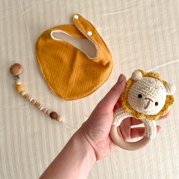 A woman's mother's hand holds a rattle with a knitted lion over the bed in which there is a cotton muslin bib and a chain with a nipple clothespin