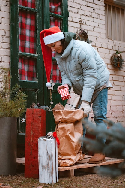 Woman in a Santa hat carries a bag of gifts from the front door of the porch of the house.