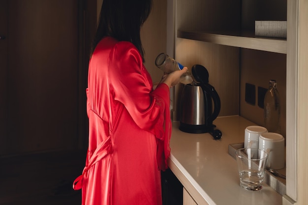 A woman in a satin nightgown pouring clean water from a bottle into a kettle in the kitchen at night