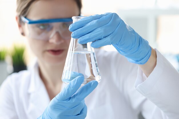 Woman scientist holding flask with transparent liquid in her hands closeup. Checking water quality and ph concept